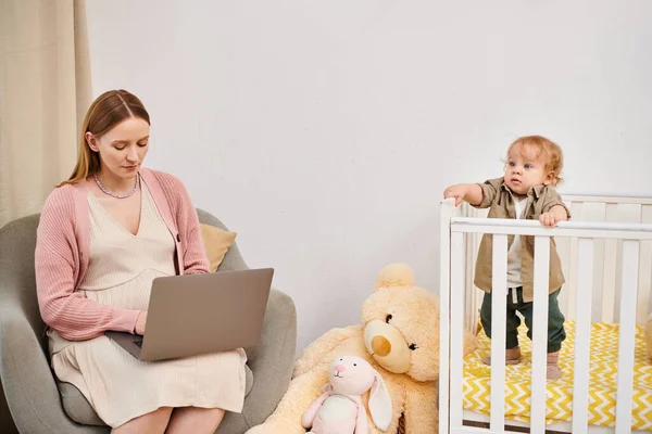 Pregnant woman working on laptop near toddler son in baby crib in nursery room, multitasking — Stock Photo