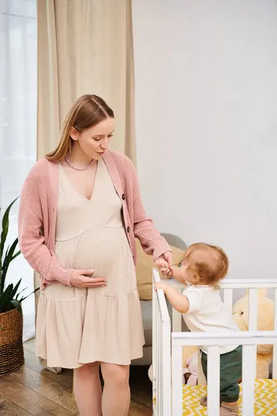 Mujer embarazada feliz cogida de la mano de su hijo pequeño de pie en la cuna en la acogedora habitación de la guardería, la maternidad - foto de stock