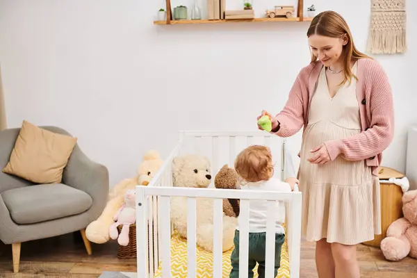 Веселая беременная женщина держит игрушку и играет с малышом сыном стоя в кроватке в детской комнате — стоковое фото
