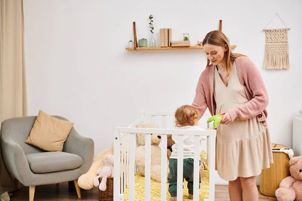 Sonriendo mujer embarazada sosteniendo juguete y jugando con su hijo pequeño de pie en la cuna en la habitación de la guardería - foto de stock