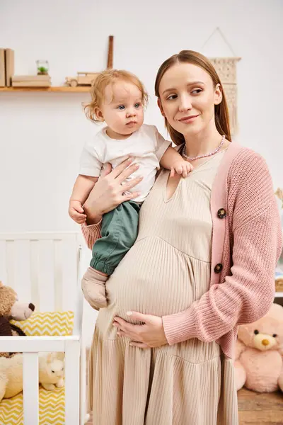 Feliz joven embarazada de pie con su hijo pequeño en la habitación de la guardería en casa, la maternidad feliz - foto de stock