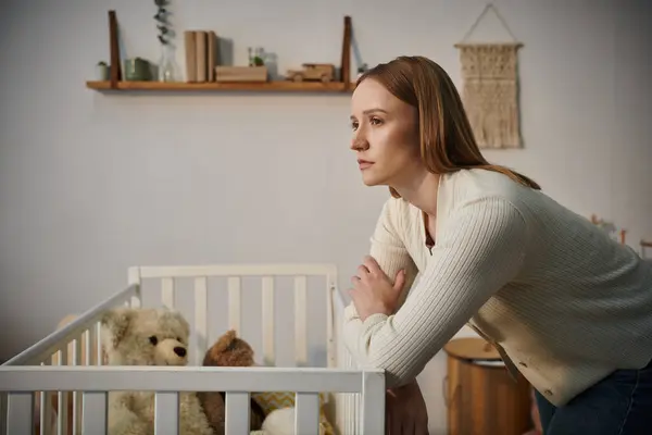 Disheartened woman standing near crib with soft toys in dark nursery room at home, grieving — Stock Photo