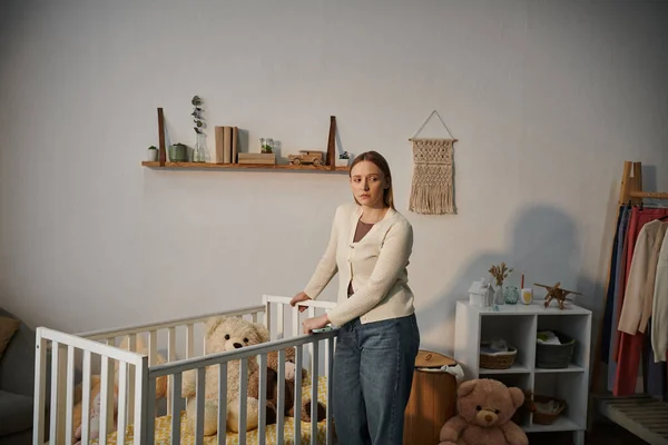 Mujer joven abrumada y solitaria de pie cerca de la cuna con juguetes suaves en la habitación de la guardería sombría en casa - foto de stock