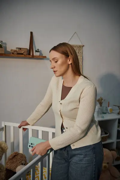 Mujer joven en duelo con teléfono inteligente de pie cerca de la cuna con juguetes blandos en la habitación de la guardería sombría en casa - foto de stock