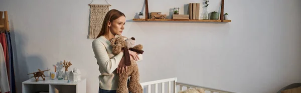 Подавленная молодая женщина с мягкой игрушкой, стоящая возле кроватки в темной детской комнате дома, баннер — стоковое фото
