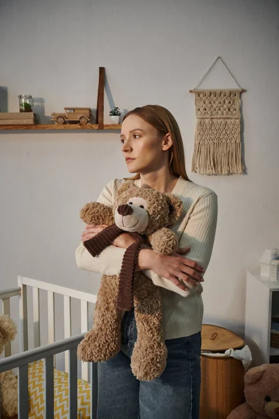 Femme au cœur lourd en deuil avec jouet doux debout près de la crèche dans la chambre de pépinière sombre à la maison — Photo de stock