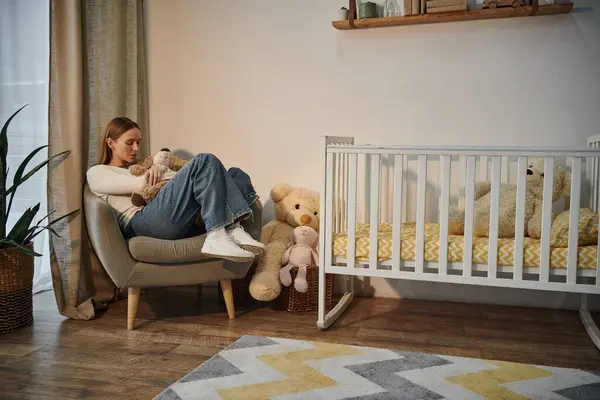 Desolate young woman with soft toy sitting in armchair near crib in bleak nursery room at home — Stock Photo