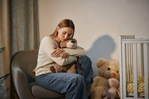 Сумна сльозова жінка з м'якою іграшкою, що сидить у кріслі біля дитячого ліжечка в похмурій кімнаті вдома — стокове фото