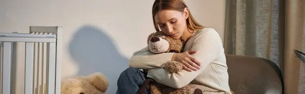 Jovem deprimida com brinquedo macio sentado em poltrona perto berço em quarto berçário sombrio, banner — Fotografia de Stock