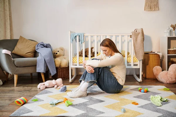 Young defeated woman sitting on floor with bowed head near toys and crib in nursey room at home — Stock Photo