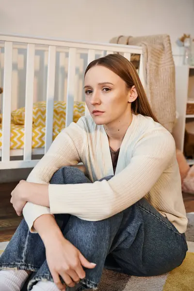 Молодая обескураженная женщина, сидящая на полу и отводящая взгляд от детской кроватки в детской комнате дома — стоковое фото