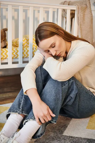 Joven mujer deprimida sentada en el suelo con los ojos cerrados cerca de la cuna en la habitación de la guardería en casa - foto de stock