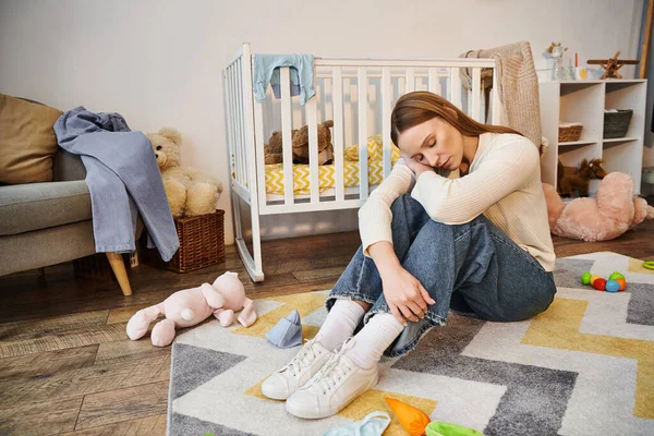 Junge hoffnungslose Frau sitzt mit geschlossenen Augen auf dem Boden in der Nähe von Babybett und Stofftieren im Kinderzimmer — Stockfoto