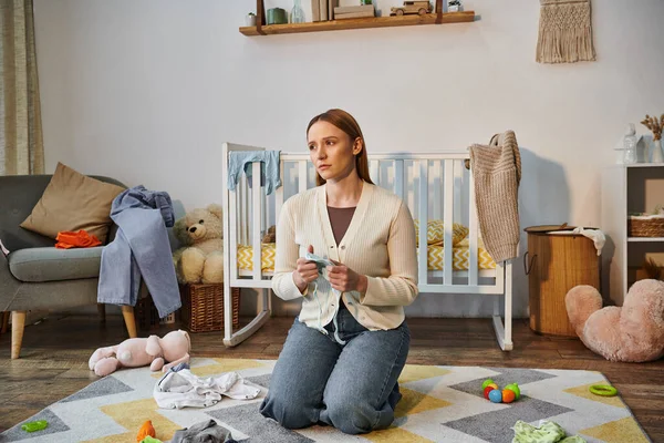 Distressed woman with baby clothes sitting on floor near crib and toys in nursery room at home — Stock Photo