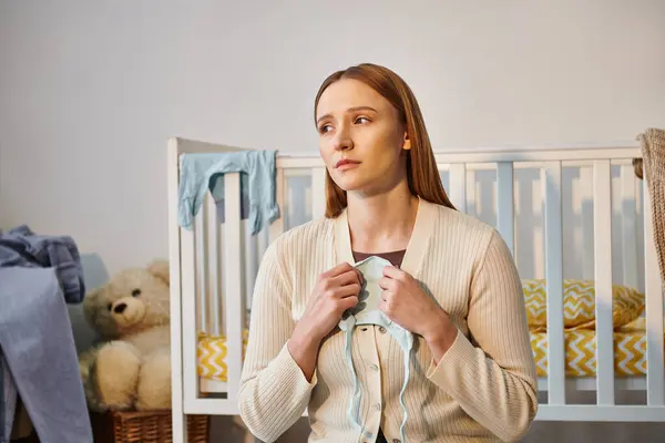 Junge trauernde Frau hält Babykleidung in der Hand und schaut in der Nähe der Krippe im Kinderzimmer weg — Stockfoto
