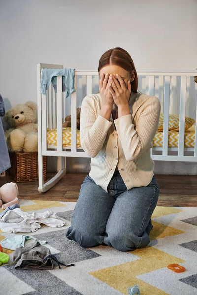 Mulher jovem e de luto chorando perto de roupas de bebê e berço com brinquedos macios no quarto de crianças — Fotografia de Stock
