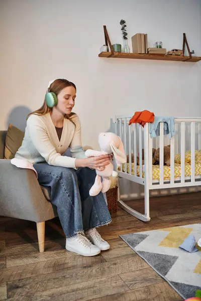 Mujer sin esperanza sosteniendo un juguete suave y escuchando música en auriculares en la habitación de la guardería oscura en casa - foto de stock