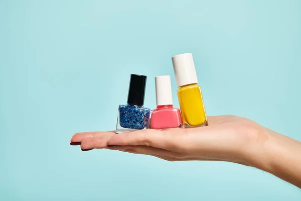 Young unknown female model holding blue pink and yellow nail polish on her hand on blue backdrop — Stock Photo