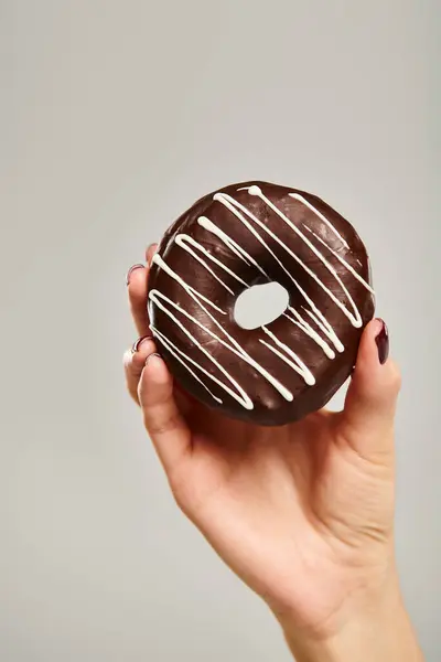 Objektfoto von Gourmet-Donut mit braunem Zuckerguss in der Hand einer unbekannten Frau auf grauem Hintergrund — Stockfoto