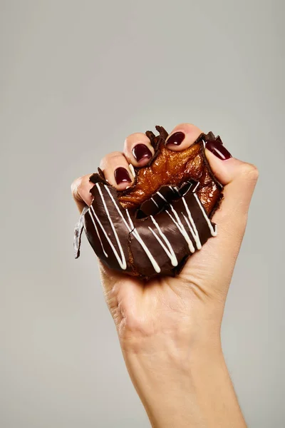 Inconnu jeune femme avec vernis à ongles serrant donut savoureux avec glaçage brun sur fond gris — Photo de stock