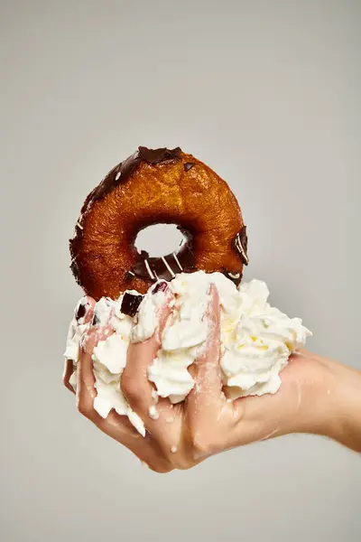 Foto objeto de delicioso donut com cobertura marrom e chantilly na mão de jovem mulher desconhecida — Fotografia de Stock