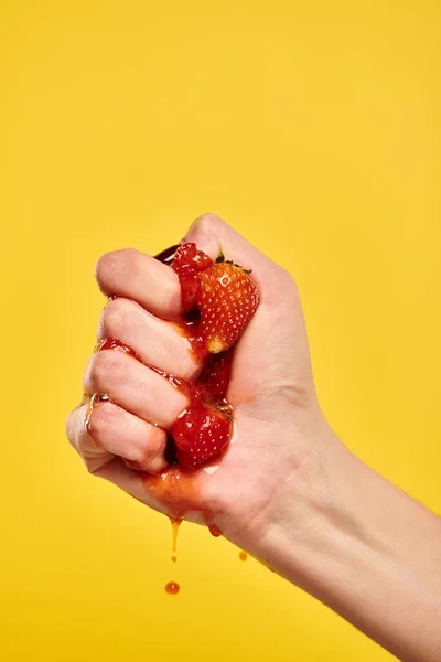Mujer joven desconocida con esmalte de uñas exprimiendo fresas frescas jugosas rojas sobre fondo amarillo - foto de stock