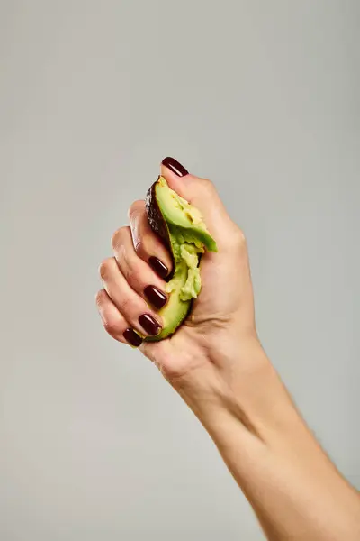 Unknown young woman with nail polish squeezing actively fresh green avocado on gray background — Stock Photo