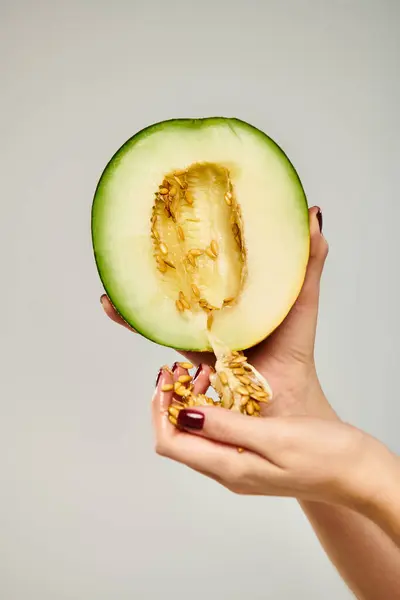 Mujer desconocida con esmalte de uñas recogiendo semillas de delicioso melón sobre fondo gris - foto de stock
