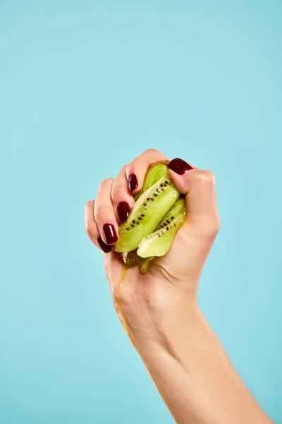 Unknown woman with nail polish squeezing green juicy kiwi in her hand on vivid blue background — Stock Photo