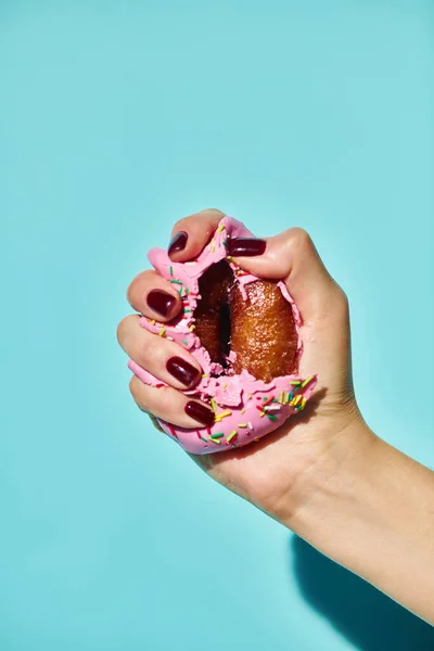 Unknown female model squeezing sweet delicious donut with pink frosting on blue background — Stock Photo