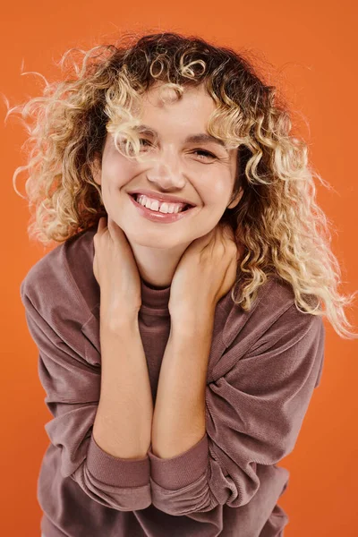 Portrait of curly happy woman in brown turtleneck smiling at camera on vibrant orange background — Stock Photo