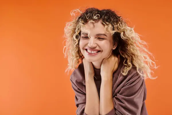 Trendy woman with wavy hair in mocha color turtleneck smiling with closed eyes on orange backdrop — Stock Photo