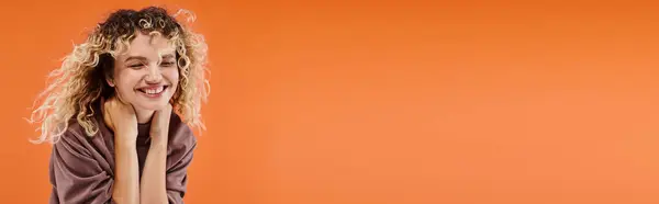 Woman with wavy hair in stylish mocha color turtleneck smiling with closed eyes on orange, banner — Stock Photo