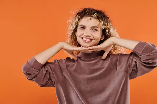 Cheerful curly woman in brown turtleneck with hands under chin looking at camera on orange — Stock Photo