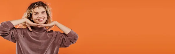 Fröhliche Frau im braunen Rollkragen mit den Händen unterm Kinn, die in die Kamera auf orangefarbenem Banner blickt — Stockfoto