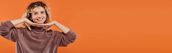 Joyful woman in brown turtleneck with hands under chin looking at camera on orange, banner — Stock Photo