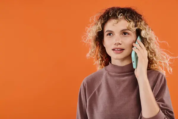 Mulher alegre com cabelo ondulado em gola alta marrom falando no telefone celular em pano de fundo laranja radiante — Fotografia de Stock