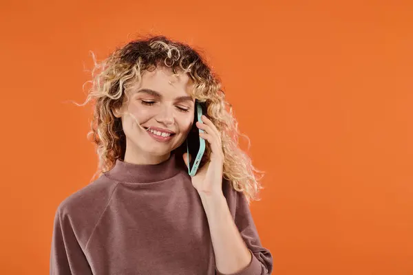 Smiling woman with curly hair in brown turtleneck talking on smartphone on radiant orange backdrop — Stock Photo