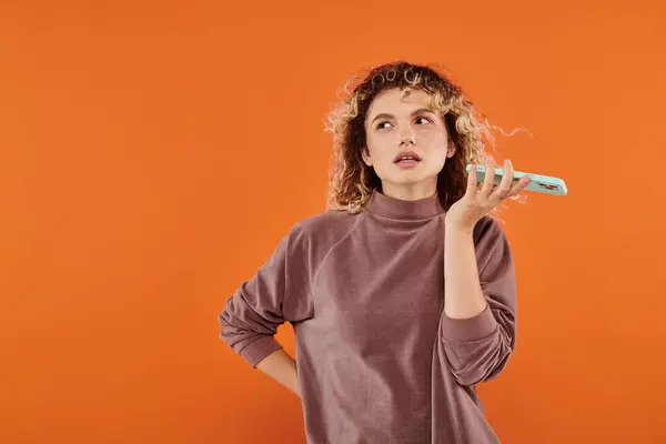 Thoughtful curly woman in mocha color turtleneck recording voice message on smartphone on orange — Stock Photo