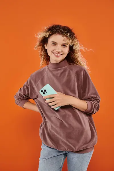 Mujer rizada feliz en cuello alto marrón sosteniendo el teléfono móvil y mirando a la cámara en el fondo naranja - foto de stock