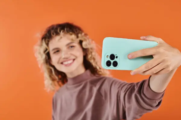Femme à la mode souriante aux cheveux ondulés prenant selfie sur mobile sur fond orange flou — Photo de stock