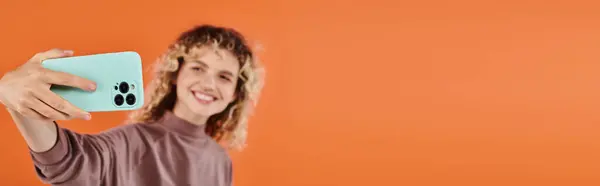 Femme à la mode souriante aux cheveux ondulés prenant selfie sur smartphone sur fond orange flou, bannière — Photo de stock