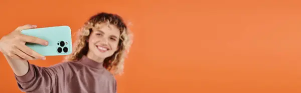 Sorrindo mulher na moda com cabelo ondulado levando selfie no smartphone em pano de fundo laranja borrado, banner — Fotografia de Stock