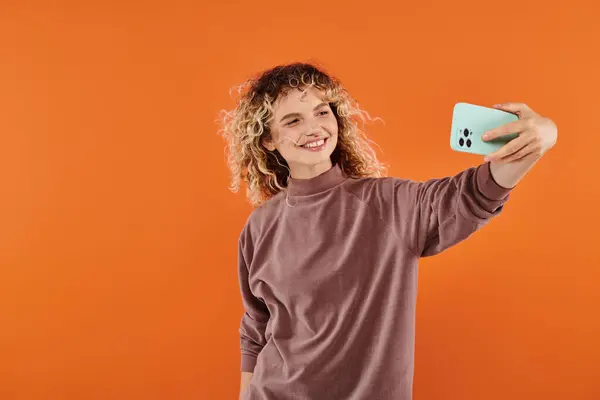Mulher elegante positivo com cabelo ondulado tomando selfie no celular no fundo laranja radiante — Fotografia de Stock