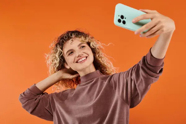 Excité femme élégante avec cheveux ondulés prendre selfie sur mobile sur fond orange radiant — Photo de stock