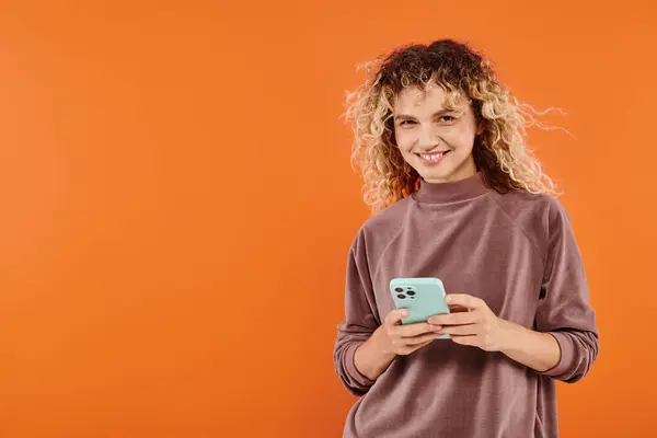 Mujer rizada encantada en la mensajería de cuello alto marrón en el teléfono móvil en el fondo del estudio naranja - foto de stock