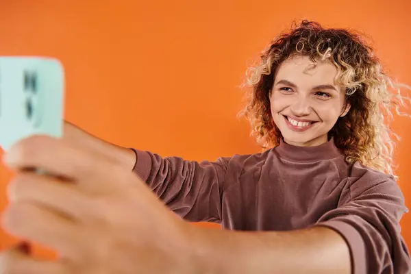 Excited woman with wavy hair taking selfie on blurred mobile phone on radiant orange backdrop — Stock Photo