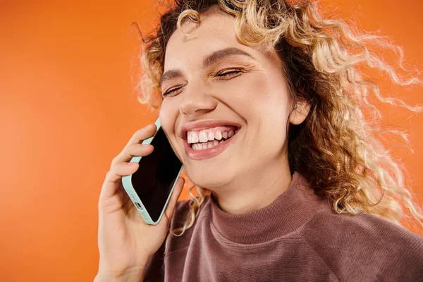 Overjoyed curly woman laughing during mobile conversation on bright orange backdrop, happiness — Stock Photo