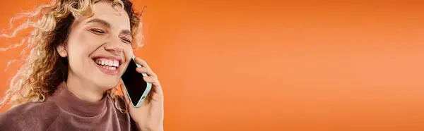 Cheerful woman with wavy hair laughing during conversation on smartphone on orange backdrop, banner — Stock Photo