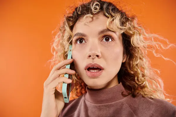 Surprised woman with curly hair and open mouth looking away while talking on smartphone on orange — Stock Photo